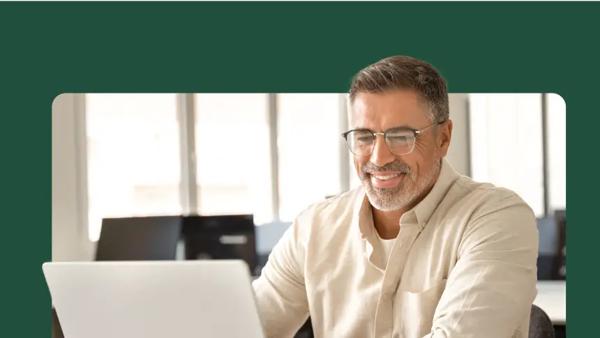 A smiling man with a beard and glasses is looking at a laptop screen.