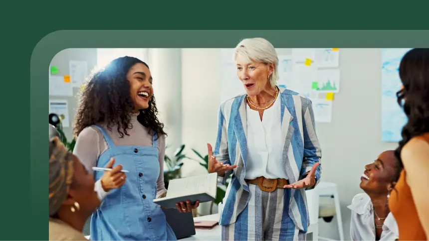Diverse group of professional women laughing and collaborating in a modern office, led by an experienced, silver-haired manager.