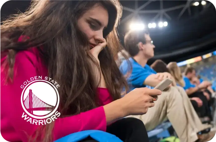 Woman looking at phone while sitting at a sports game, next to logo of Golden State Warriors