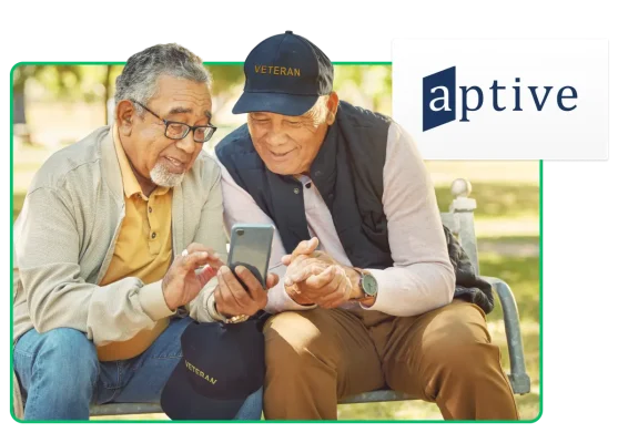 Two men with baseball caps that say "Veteran" look at a cell phone