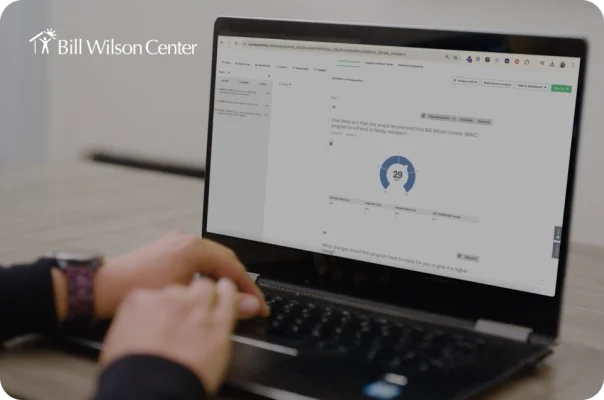 A person typing on a laptop showing a Bill Wilson Center program survey dashboard with an NPS gauge.