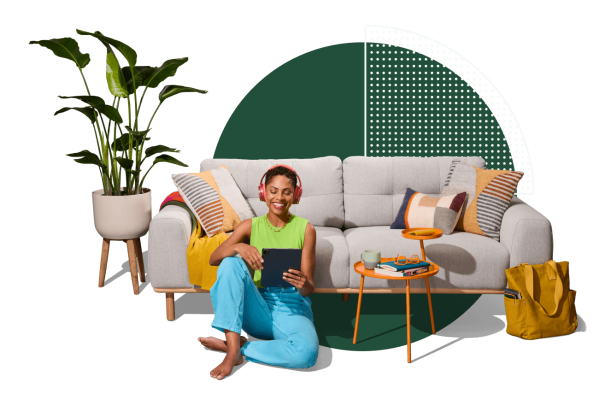 Vrouw zit op de grond met een koptelefoon en kijkt naar een tablet, ze leunt tegen een bank met daarnaast een plant en een bijzettafel