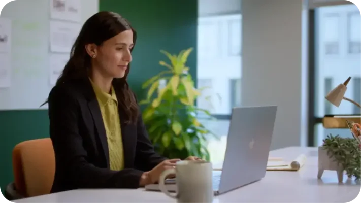 A woman in a black blazer and yellow shirt smiling while working on a laptop at her office desk