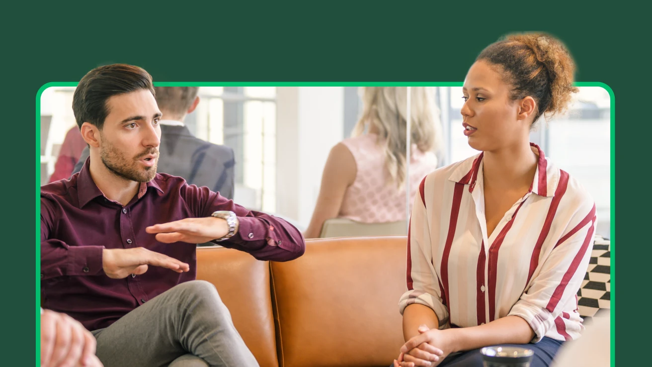 Two people in business casual clothing sitting on a couch and having a discussion