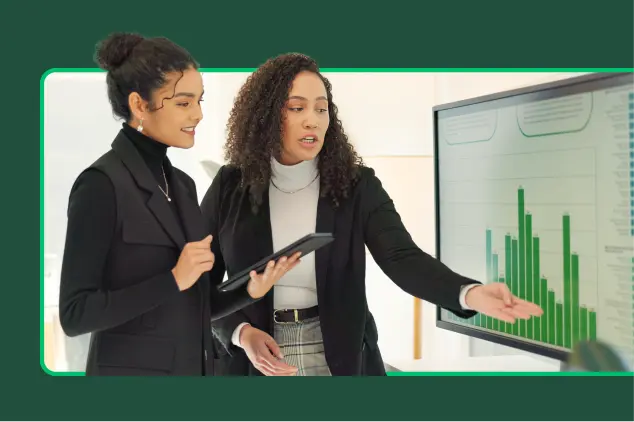 Woman doing a presentation and pointing to a bar graph on the screen, with another woman listening and taking notes
