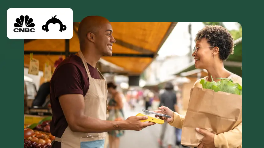 A small business vendor at a market smiling while a customer pays with a mobile device, featuring the CNBC and SurveyMonkey logos.