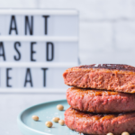 Tre hamburger impilati su un piatto con alle spalle un cartello con la scritta "PLANT BASED MEAT" (carne vegetale)
