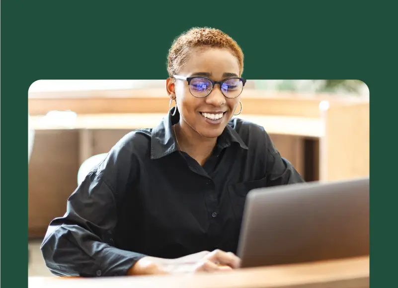 A woman with short, blonde hair and glasses is smiling while looking at a laptop screen.